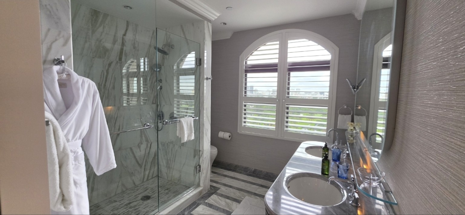 Marble bathroom with double sinks, glass shower, robes, and plantation shutters