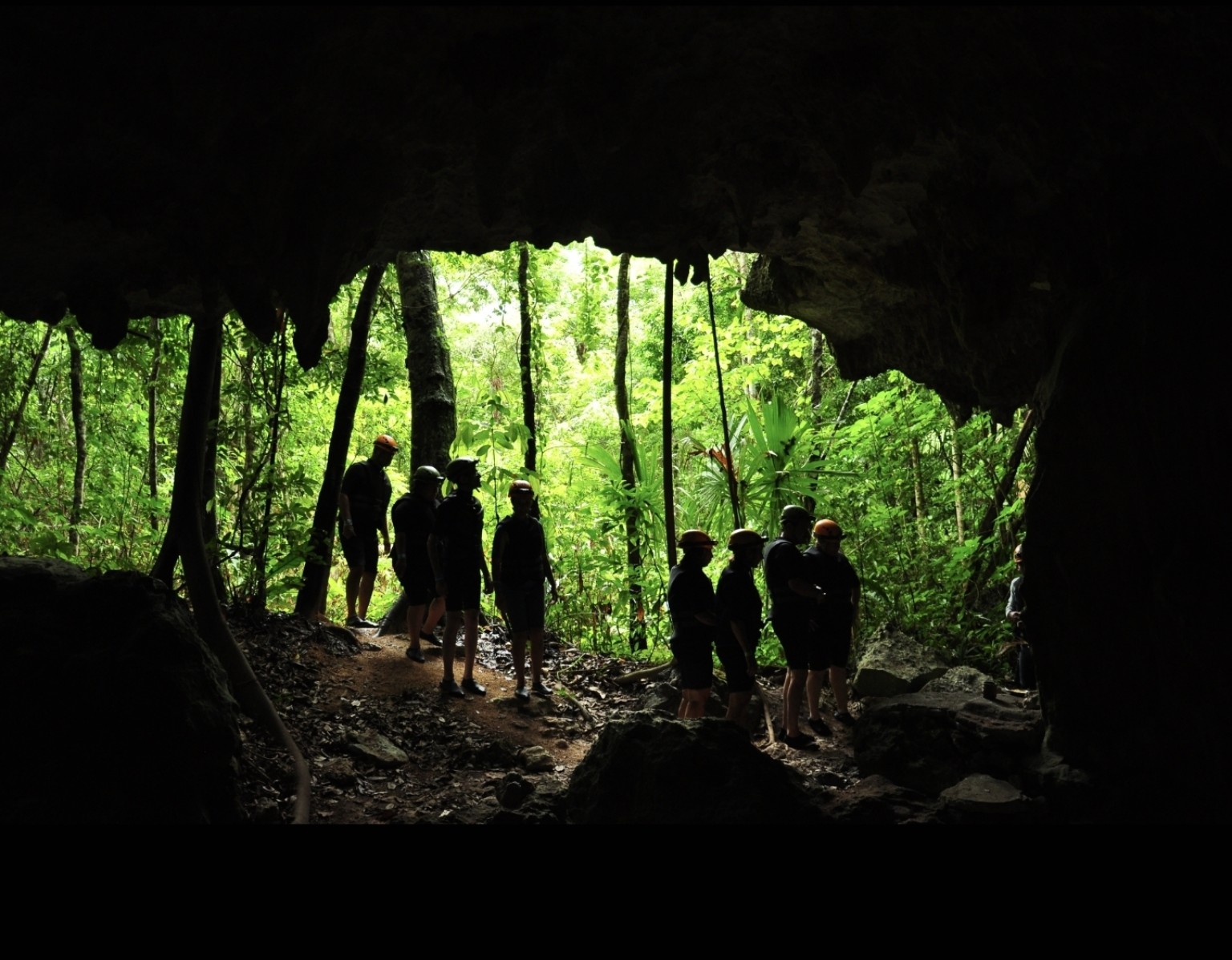 A guided Río Secreto group silhouetted at a cave opening surrounded by bright green jungle