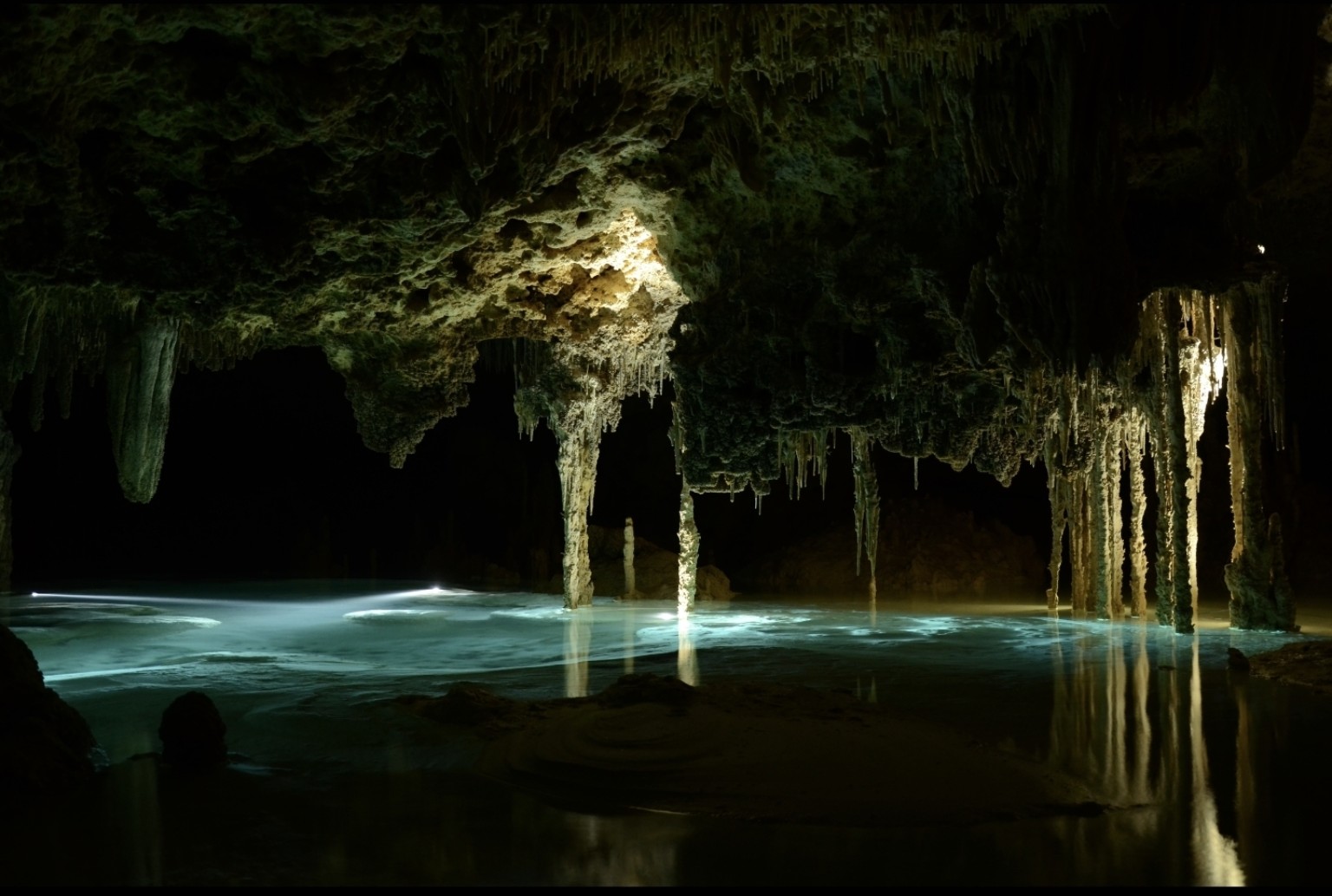 Dark Río Secreto chamber with illuminated turquoise water and long stalactites